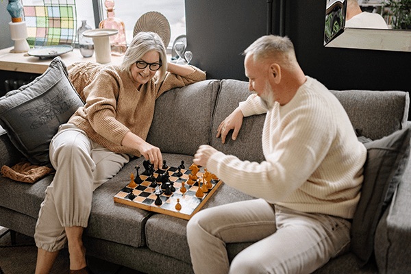 An older woman and man sit on a gray couch, smiling and playing chess together in a cozy, well-lit living room at MorningStar Senior Living Kirkland. The atmosphere is relaxed and comfortable, capturing the joy of retirement living.